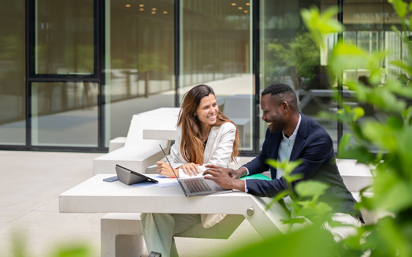 A man and a woman sitting outside talking, working on a tablet and laptop