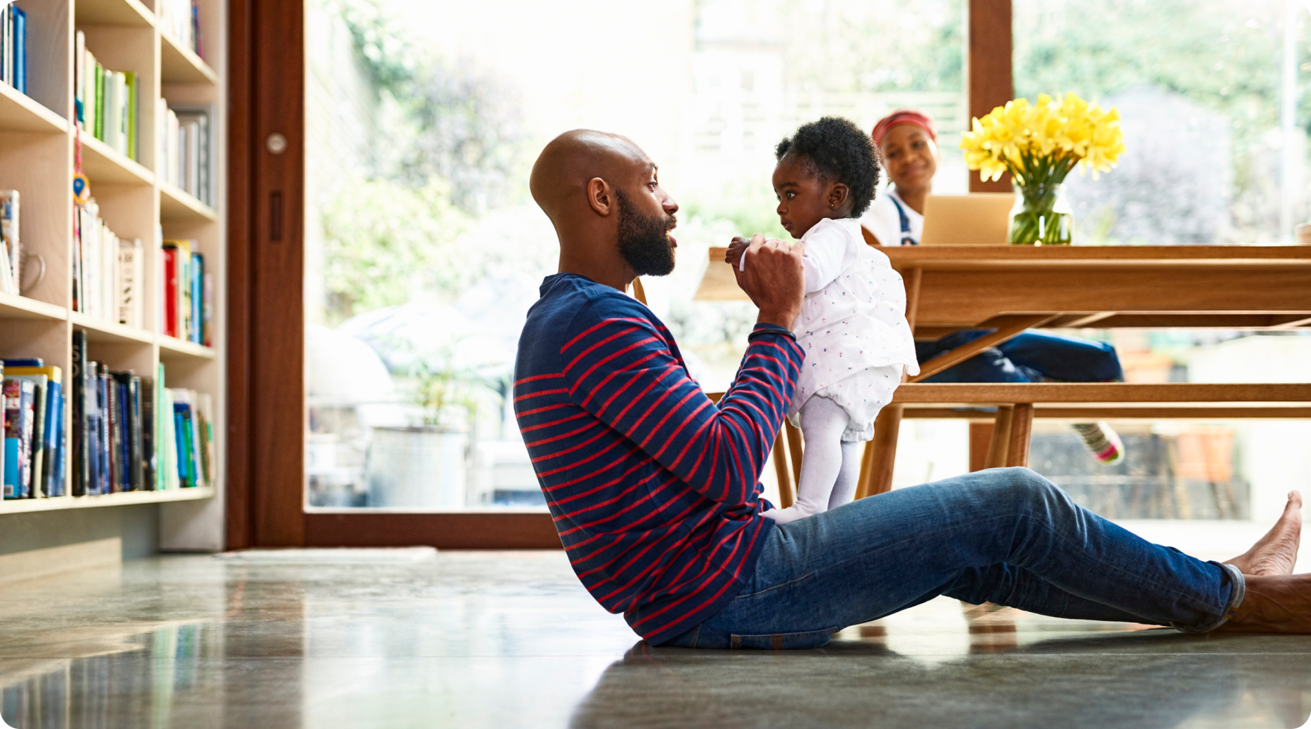 Image of man sitting on ground with toddler girl standing on top of his legs, as woman watches in the background